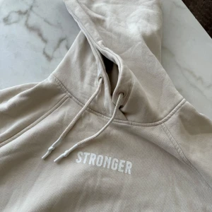 Beige hoodie från Stronger - Säljer en snygg beige hoodie från Stronger med texten 'STRONGER' tryckt på bröstet. Hoodien har en klassisk känguruficka och justerbar dragsko i huvan. Perfekt för en avslappnad stil.