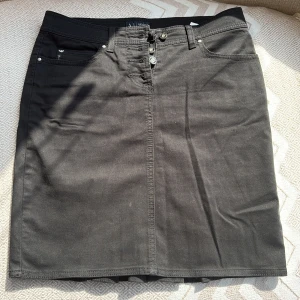 Svart jeanskjol från Armani Jeans - Snygg svart jeanskjol från Armani Jeans med knappar framtill och klassiska femficksdetaljer. Kjolen har en stilren design med plisserad detalj baktill som ger en unik touch. Perfekt för en trendig och avslappnad look.