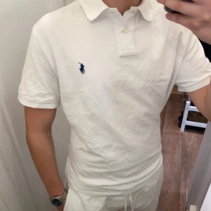 Polo Ralph Lauren (Vit piké) - Säljer en klassisk vit piké från Polo Ralph Lauren med korta ärmar och den klassiska loggan i blått på bröstet. Perfekt för en stilren och avslappnad look. Den har inga häl eller fläckar utan den är i underbar skick och redo för en ny ägare! Den är size XL men passar L!