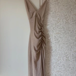 Champagnefärgad glittrig långklänning - Säljer en  champagnefärgad långklänning med glittrigt tyg och v-ringning. Klänningen har smala axelband och snygg rynkdetalj längst sidan. Bara använt klänningen vid 1 tillfälle innan.