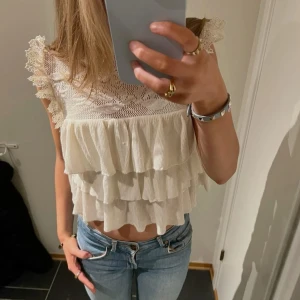 Vit volangblus med spetsdetaljer från Zara - Supersöt vit blus med flera volanger och spetsdetaljer på axlarna. Blusen har ett vackert blommönster upptill och är ärmlös. Perfekt att styla med jeans eller kjol för en trendig look.              Första bilden är lånad! 