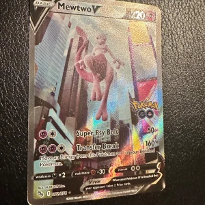 Mewtwo V Pokémonkort - Säljer ett holografiskt Mewtwo V-kort från Pokémon GO-serien. Kortet har en färgstark och glansig framsida med Mewtwo i en stadsmiljö. Det är ett basic V-kort med attacker som Super Psy Bolt och Transfer Break.