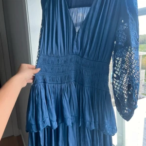 Blå klänning med spetsdetaljer från Maje - Supersnygg blå klänning från Maje med v-ringning och långa ärmar i spets. Klänningen har smockad midja och volang nertill, vilket ger en feminin siluett. Perfekt för dig som vill sticka ut med unika detaljer och elegant design.