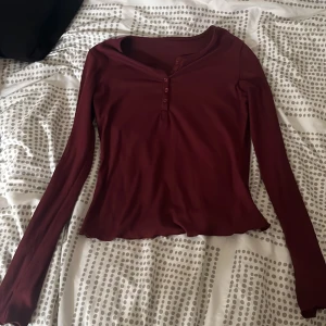 Vinröd långärmad topp från Dazy - Säljer en vinröd långärmad topp från Dazy med knappar framtill och lätt vågig kant nertill. Toppen har en smickrande v-ringning och är i ett mjukt, stretchigt material. Perfekt att styla med jeans eller kjol.