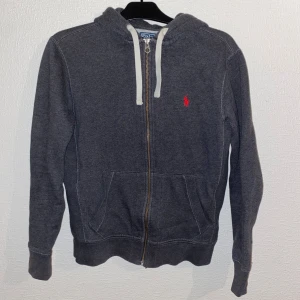 Grå hoodie från Polo Ralph Lauren - Säljer en stilren grå hoodie från Polo Ralph Lauren med dragkedja och det klassiska röda logotypbroderiet på bröstet. Tröjan har långa ärmar och en praktisk huva med snörning. Perfekt för en avslappnad look!