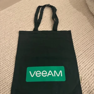 mörkgrön tygkasse med Veeam-tryck - Säljer en mörk grön tygkasse med grönt och vitt Veeam-tryck på framsidan. Väskan har två långa handtag och är tillverkad i ekologisk bomull. Perfekt för att bära böcker, mat eller andra vardagsprylar.