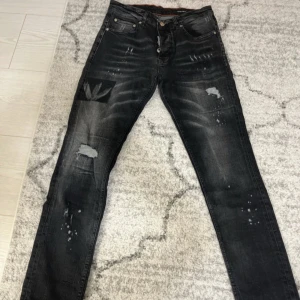 Svarta jeans med slitningar från Dsquared2 - Säljer ett par svarta jeans från Dsquared2 med coola slitningar och detaljer. Jeansen har en normal passform, klassisk femficksdesign och knappgylf. Snygga slitna partier och unika tryckdetaljer på ena benet.