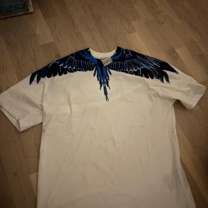 Vit t-shirt med blått vingmönster från Marcelo Burlon - Säljer en vit t-shirt från Marcelo Burlon med ett snyggt blått och svart vingmönster över axlar och hals. T-shirten har normal passform och korta ärmar. Perfekt för dig som vill sticka ut med en unik design.