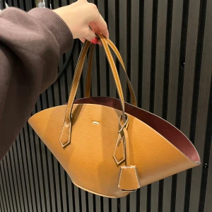 Brun handväska i skinn med unik form - Säljer en snygg brun handväska i skinn med en cool, vid form och dubbla handtag. Väskan har en innerpåse med dragsko och kontrasterande mörkröd insida. Perfekt för dig som vill sticka ut med din accessoar. Helt oanvänd väska aldrig använt 