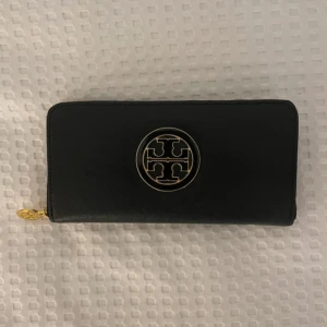 Svart plånbok från Tory Burch - Stilren svart plånbok från Tory Burch med guldfärgad logga och dragkedja. Plånboken har flera kortfack och ett innerfack med dragkedja. Perfekt för dig som vill ha en klassisk och elegant accessoar. Dock är den lite smutsig inuti!!!