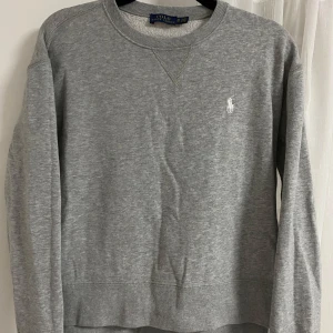 Grå sweatshirt från Polo Ralph Lauren - Säljer en klassisk grå sweatshirt från Polo Ralph Lauren med rund halsringning och vit broderad logga på bröstet. Tröjan har långa ärmar och är perfekt för en avslappnad stil.