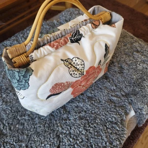 Blommig handväska med bambuhandtag - Charmig handväska med ett blommigt mönster i flera färger och bambuhandtag. Väskan har en mjuk, vadderad insida och en unik design som ger en bohemisk känsla. Perfekt för att ge din outfit en färgglad touch.