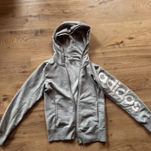 Grå hoodie från Adidas Essentials - Grå hoodie från Adidas Essentials med dragkedja och huva. Stor Adidas-logga i vitt längs ena ärmen. Perfekt för en sportig och avslappnad stil. Mjuk bomullsblandning och två fickor framtill.