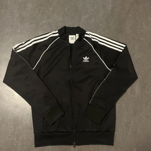Svart Adidas Zip-up kofta - Säljer en svart träningsjacka från Adidas med de klassiska tre ränderna längs ärmarna. Jackan har dragkedja framtill och Adidas-loggan på bröstet. Perfekt för en sportig look eller träning.