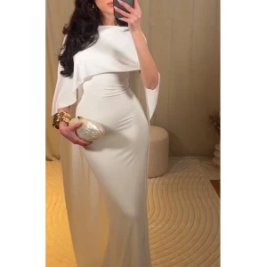 Vit långklänning med cape - Elegant vit långklänning med en stilren cape-design. Klänningen har en figurnära passform och är perfekt för formella tillfällen. Den är axelbandslös och har en vacker drapering över axlarna.