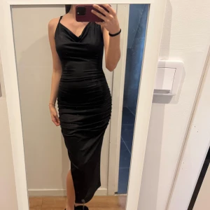 Svart axelbandslös klänning - Elegant svart klänning med smala axelband och en smickrande drapering framtill. Klänningen har en slits som ger en extra touch av stil. Perfekt för en kväll ute eller en speciell tillställning.