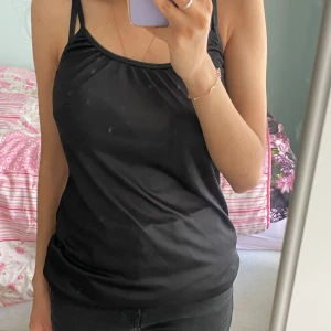 Svart linne med smala axelband - Säljer en svart linne med smala axelband och enkel, stilren design. Linnet har en rak passform och är tillverkad i ett mjukt, glansigt material som ger en elegant känsla. Perfekt för dig som gillar minimalistisk stil.