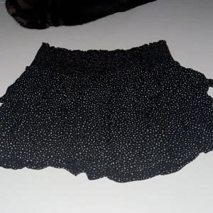 Svart prickig kjol från Kappahl - Säljer en svart kortkjol från Kappahl med vita prickar. Kjolen har en elastisk midja för en bekväm passform. Perfekt för en stilren look.