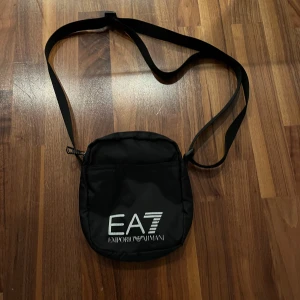  Emporio Armani EA7 - Snygg svart axelväska från Emporio Armani med justerbar rem och logotypen EA7 framtill. Perfekt för att bära med sig det viktigaste på ett stilfullt sätt.