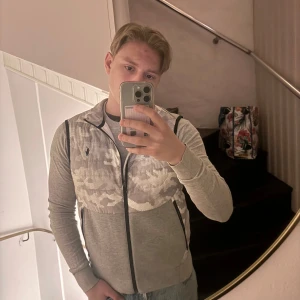 Grå väst från Polo Ralph Lauren - Säljer en snygg grå väst från Polo Ralph Lauren med ett subtilt camouflage-mönster. Västen har dragkedja och är perfekt för en stilren look. Passar bra till både jeans och chinos. Sitter som S