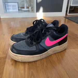 Nike Air Force med rosa detaljer - Säljer ett par Nike Air Force sneakers i svart med en iögonfallande rosa swoosh. Skorna har en klassisk design med snörning och en robust sula. Perfekta för dig som vill ha en stilren men ändå färgglad look. Missfärgade där det ska vara vitt, är numera gult. Har inte testat att ta bort så går möjligtvis bort 😊passar 38-39
