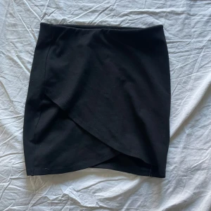 Svart omlottkjol från Gina Tricot - Snygg svart omlottkjol från Gina Tricot i en kort modell. Kjolen har en stilren design med omlott-detalj framtill som ger en elegant touch. Perfekt för att skapa en chic look.