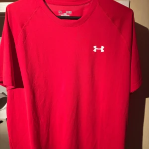Röd t-shirt från Under Armour - Säljer en röd t-shirt från Under Armour med klassisk logga på bröstet. T-shirten är kortärmad och har en loose fit, perfekt för träning eller vardag. Materialet är syntet och modellen är från HeatGear-serien.