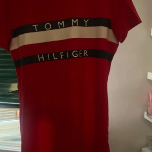 Röd t-shirtklänning från Tommy Hilfiger - Röd t-shirtklänning från Tommy Hilfiger med vita och svarta ränder samt logga över bröstet. Klassisk rund halsringning och korta ärmar. Perfekt för en avslappnad och sportig look.
