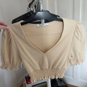 Beige croppad topp från NA-KD - Säljer en beige croppad topp från NA-KD med puffärmar och v-ringning. Toppen har resår i midjan och vid ärmsluten för en snygg passform. Perfekt för vår och sommar!