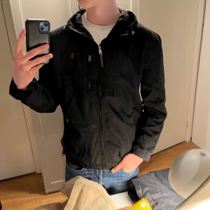 Svart vindjacka från Polo Ralph Lauren - Svart vindjacka från Polo Ralph Lauren med dragkedja framtill och liten röd broderad logga på bröstet. Jackan har huva, flera fickor med dragkedja och en klassisk passform. Perfekt för blåsiga dagar.
