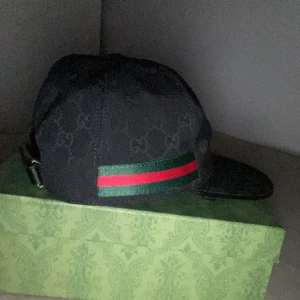 Svart keps med grönt och rött band - Snygg svart keps med diskret mönster och ett grönt och rött band. Justerbar rem baktill för perfekt passform. Perfekt för en stilren look.