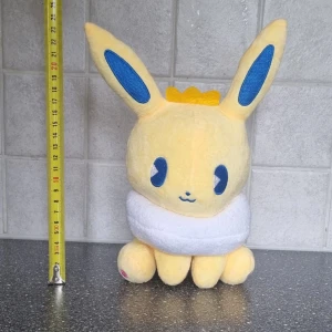 Gul Eevee, pokemonfigur, gosedjur, plush - Söt gul Eevee plush med stora blå ögon och öron. Den har en vit krage och en liten gul krona på huvudet. Perfekt för kramar och lek!