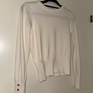 Vit tröja med knappar - Säljer en stilren vit tröja med långa ärmar och dekorativa knappar vid ärmsluten. Tröjan har en rund halsringning och ribbade detaljer vid ärmslut och nederkant. Perfekt för en elegant och enkel look.