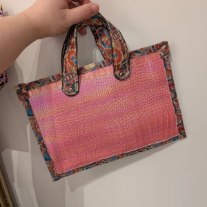 Färgglad handväska med krokodilmönster - Unik handväska med ett färgglatt paisleymönster och krokodilmönstrad yta i rosa och lila toner. Väskan har ett rymligt fack och bärhandtag i matchande mönster. Perfekt för att ge din outfit en färgklick!