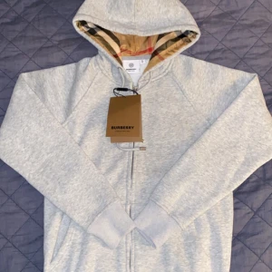 Grå hoodie från Burberry - Säljer en stilren grå hoodie från Burberry med dragkedja och klassiskt rutigt foder i huvan. Perfekt för en avslappnad och trendig look. 🧥det står S men är mer som L i Barn storlek passar folk som är runt 165 (A-Cop) 