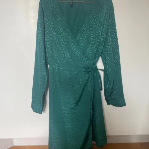 Grön omlottklänning från Vero Moda - Snygg grön omlottklänning från Vero Moda med  mönster och knytband i midjan. Klänningen har långa ärmar och en v-ringad hals💚