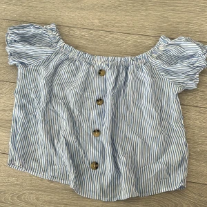 Blå- och vitrandig offshoulder-blus - Säljer en söt blå- och vitrandig offshoulder-blus med knappar framtill. Blusen har korta puffärmar och en luftig passform, perfekt för varma sommardagar. Den elastiska halsringningen ger en bekväm och flexibel passform.