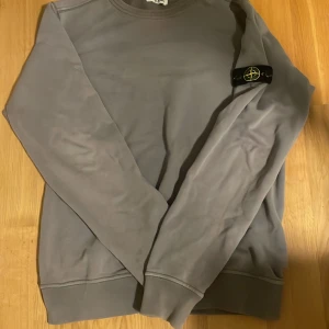 Grå sweatshirt från Stone Island - Säljer en stilren grå sweatshirt från Stone Island med deras ikoniska märke på ärmen. Tröjan har långa ärmar och en klassisk rund halsringning. Perfekt för en avslappnad stil.