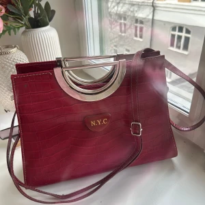 Röd axelväska med krokodilmönster - Snygg röd axelväska med krokodilmönster och silverfärgade handtag. Väskan har en N.Y.C-platta och en praktisk dragkedja upptill. Axelremmen är avtagbar och det finns innerfickor. Den är aldrig använd så i väldigt nyskick.