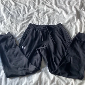 Svarta mjukisbyxor från Under Armour - Säljer ett par svarta mjukisbyxor från Under Armour med elastisk midja och snörning. Byxorna har en lös passform och är perfekta för träning eller avslappning. De har dragkedjefickor och en liten logga på benet.