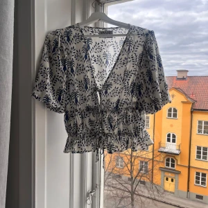 Vit och blå blus från Aware  - Säljer en söt blus från Aware i vitt med blått mönster. Blusen har korta ärmar och en knytning framtill. Perfekt för vår och sommar!
