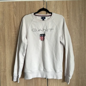 Vit GANT tröja  - Säljer en stilren vit sweatshirt från GANT med deras ikoniska logga och emblem på bröstet. Tröjan har långa ärmar och ribbade muddar vid ärmslut och nederkant. Perfekt för en avslappnad look.