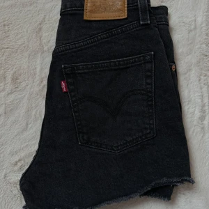 Svarta jeansshorts från Levi's - Snygga svarta jeansshorts från Levi's med fransiga kanter och klassisk femficksdesign. Modellen är ”ribcage shorts” i storlek W25. Perfekta sommarens varma väder, solsemester eller vid stranden. 