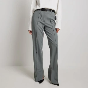 Grå kostymbyxor med slits - Det här kostymbyxan har en hög midja, en stängning med dragkedja, hake och knapp, en fuskbakficka, sidofickor, en veckad detalj, rakt ben och två sidoslitsar på varje sida.