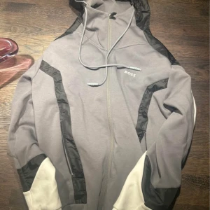 Grå och svart hoodie från Boss - Säljer en stilren grå och svart hoodie från Boss. Den har en dragkedja framtill och justerbar huva med snörning. Perfekt för en avslappnad look. Köpt för 3000kr