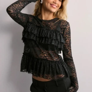 Svart spetsblus med volanger - Elegant svart spetsblus med volanger och knytning i ryggen. Blusen har ett blommigt mönster och långa ärmar, perfekt för en stilfull look.