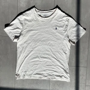 Vit t-shirt från Polo Ralph Lauren - Klassisk och stilren vit T-shirt från Ralph Lauren. Tidlös design med den ikoniska loggan på bröstet. T-shirten är i bra skick, lätt använd. Perfekt för både vardag och fritid! 	•	Storlek: S (Barn XL) – passar ungefär som en liten vuxenstorlek 	•	Färg: Vit 	•	Skick: Bra – inga hål eller stora fläckar 	•	Mjukt och bekvämt material