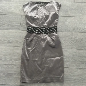 Grå sidenklänning med paljettbälte - Elegant grå klänning i siden med ett iögonfallande svart och silver paljettbälte i midjan. Klänningen har en kort ärm och en figurnära passform som ger en sofistikerad look.