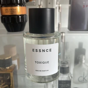 Essnce Toxique Eau de Parfum - Säljer en elegant flaska Essnce Toxique Eau de Parfum. Flaskan är genomskinlig med en minimalistisk vit etikett och svart text. Den har en svart spraykork och innehåller en doft med noter av citrus och blommor. Perfekt för den som söker en unik och sofistikerad doft.