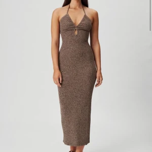 Brun stickad adoore klänning  - Snygg brun stickad klänning med halterneck och en liten öppning framtill. Klänningen är lång och har en åtsittande passform som framhäver figuren. Perfekt för en stilren look.